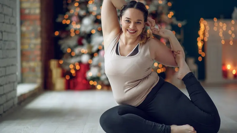 An image of a happy woman in a yoga pose, sitting on the floor of her living room loading=