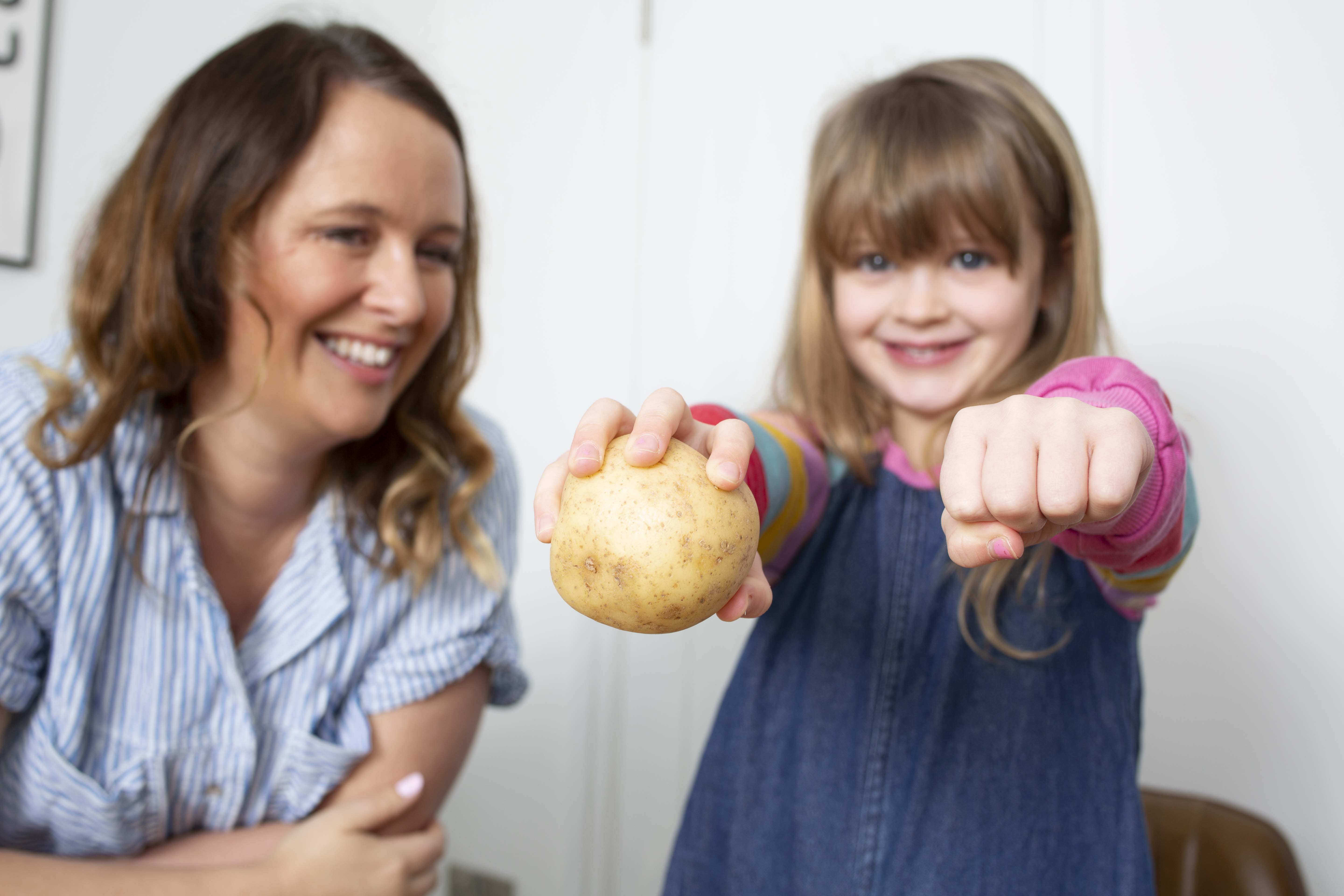A smiling woman and her young daughter. The daughter is holding a potato in one hand and her fist outstretched, to compare the size of the potato and her hand. loading=