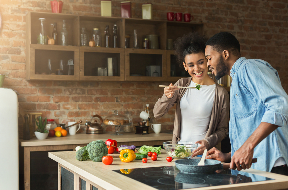A husband and wife tasting food from a spoon in their kitchen. The counter is covered with colourful vegetables, such as peppers. loading=