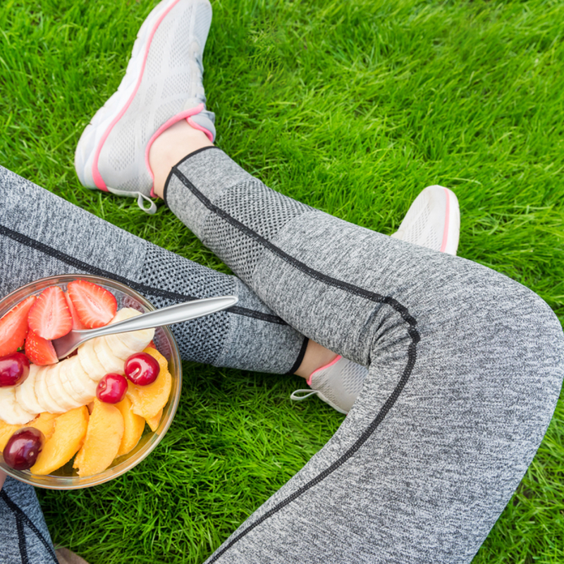 An adult wearing activewear sitting on the grass eating a fruit salad